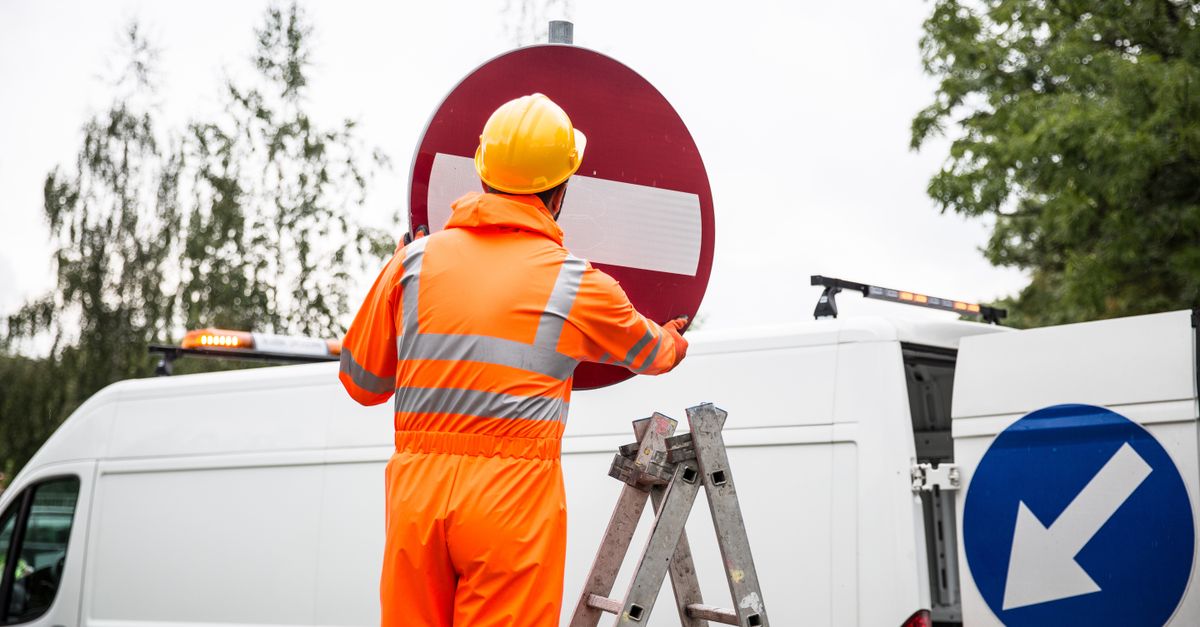 A road worker in orange uniform adjusting a traffic sign on a ladder by a van.