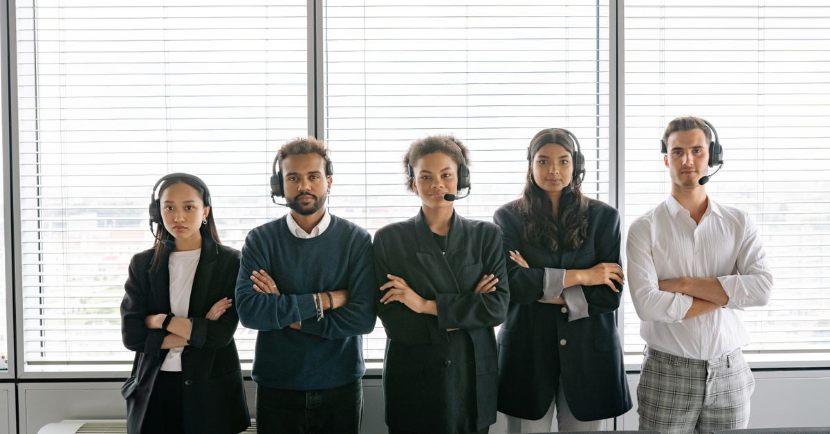 A diverse group of call center agents standing with arms crossed, wearing headsets in an office.