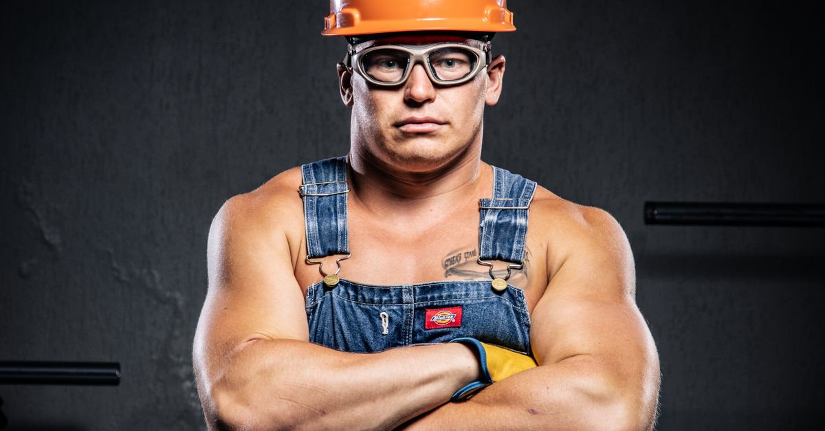 Portrait of a muscular construction worker in an orange hard hat and goggles with arms crossed.