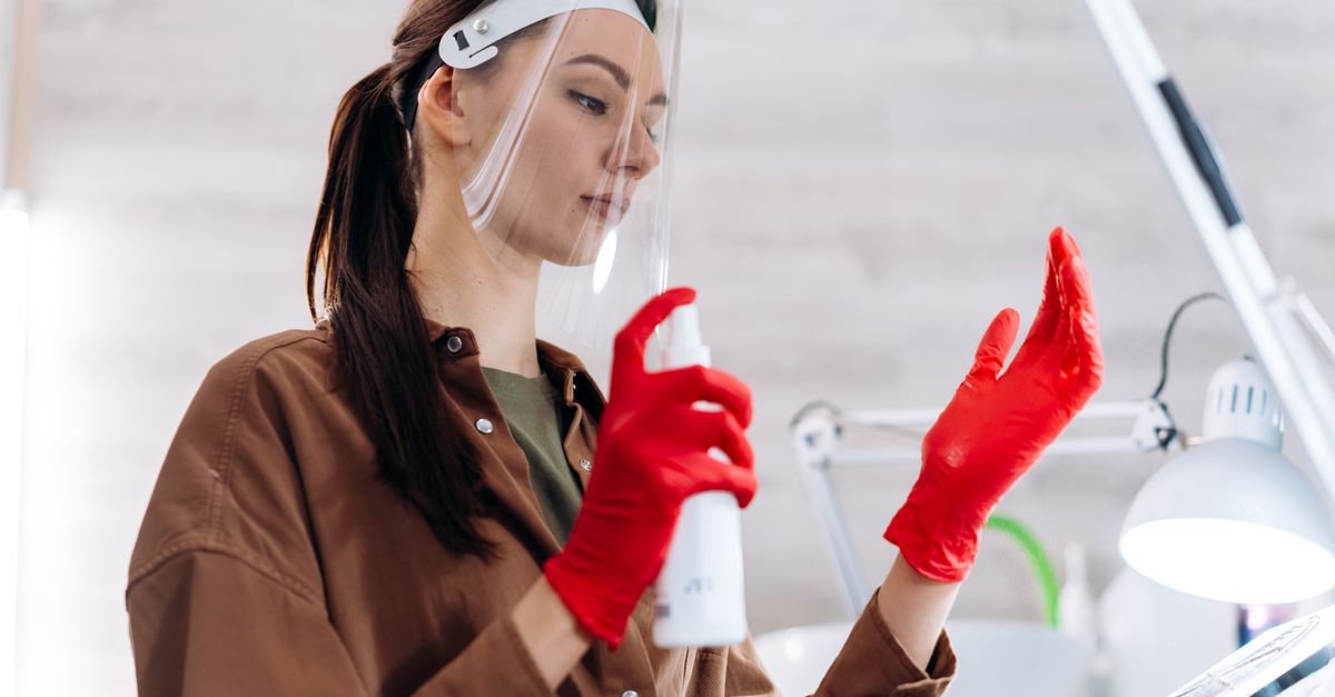 Manicurist wearing a face shield and red gloves in a salon, holding a spray bottle as part of safety measures.