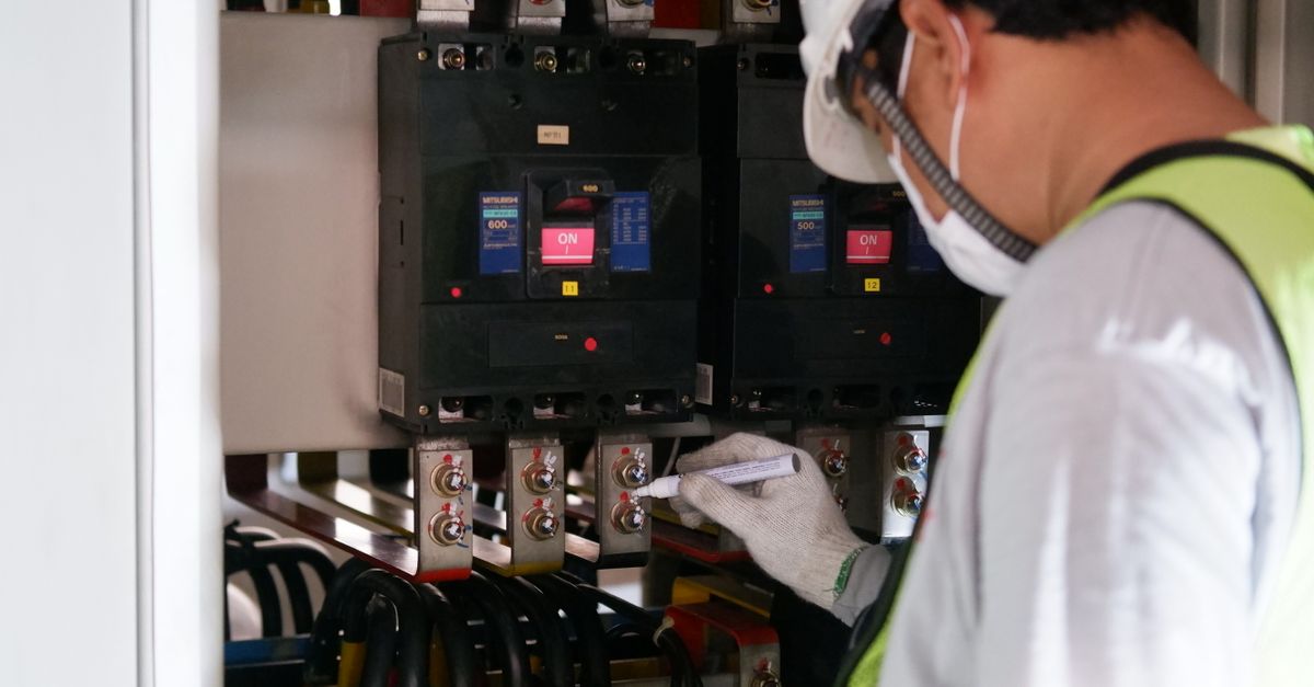 Technician wearing safety gear inspecting electrical panels in an industrial setting.