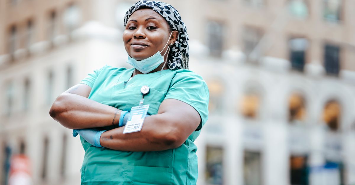 Low angle of confident African American nurse in gloves and mask standing with crossed arms and looking at camera