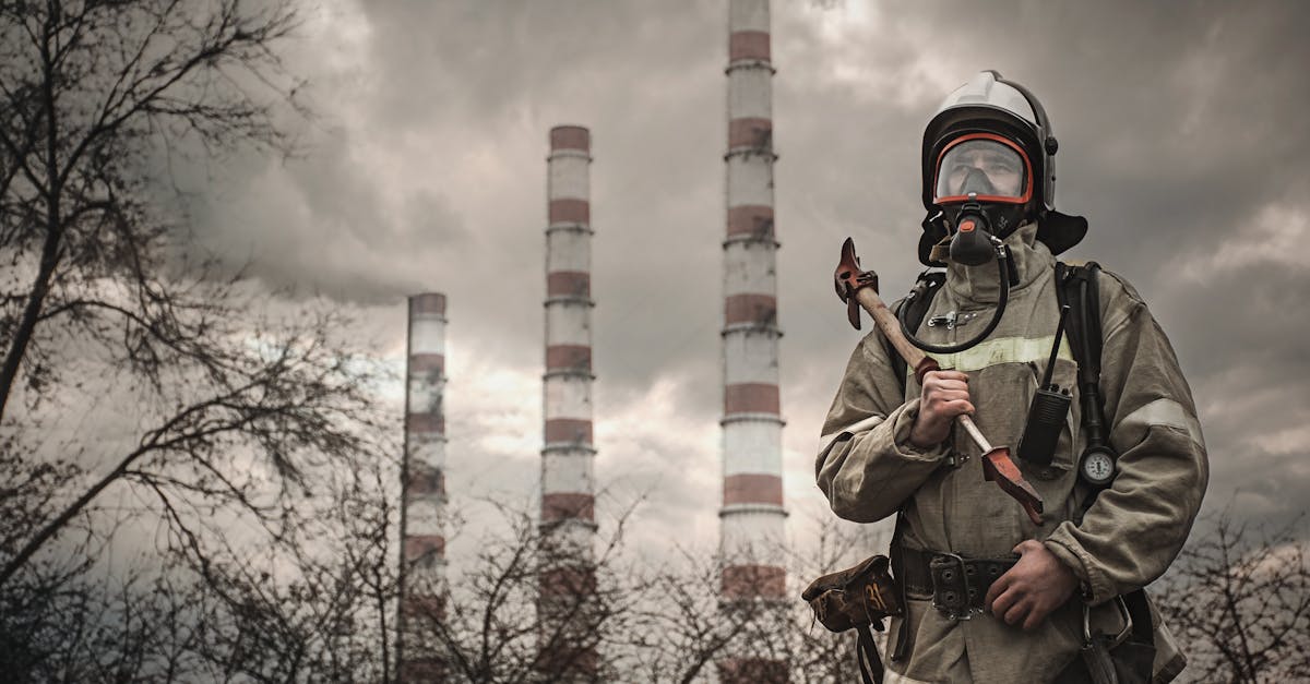 Firefighter wearing protective gear standing in front of industrial smokestacks under cloudy skies.