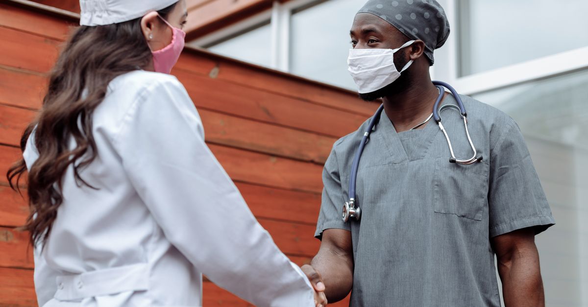Two healthcare workers in face masks shaking hands outdoors, promoting teamwork and collaboration.