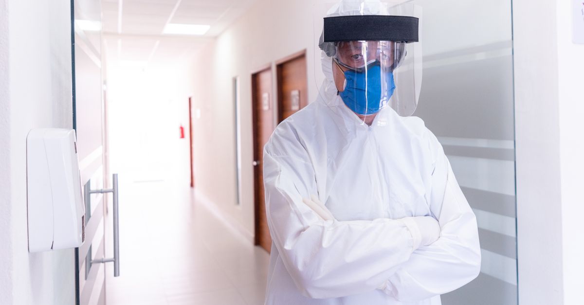Medical professional in full PPE gear stands in a hospital corridor, wearing face mask and shield.