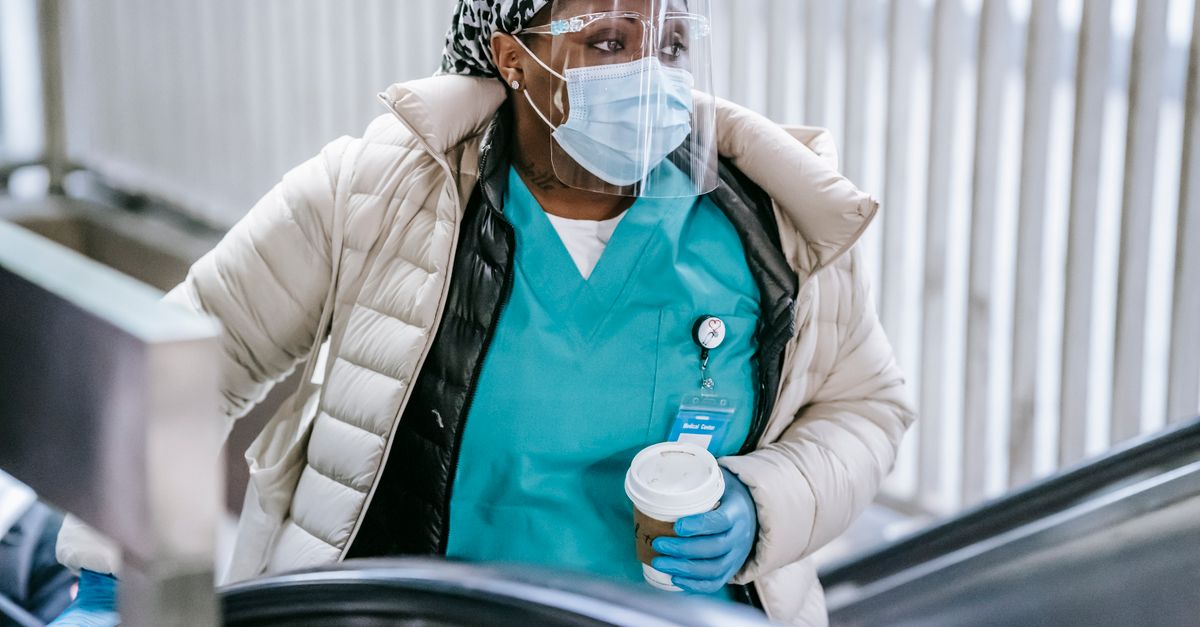 Plump ethnic female doctor in face shield with hot drink to go looking away on escalator