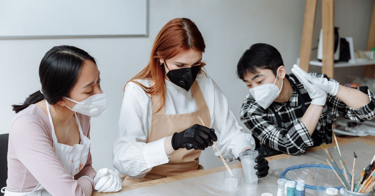Three diverse artists wearing masks and gloves experiment with colors in a creative studio environment.