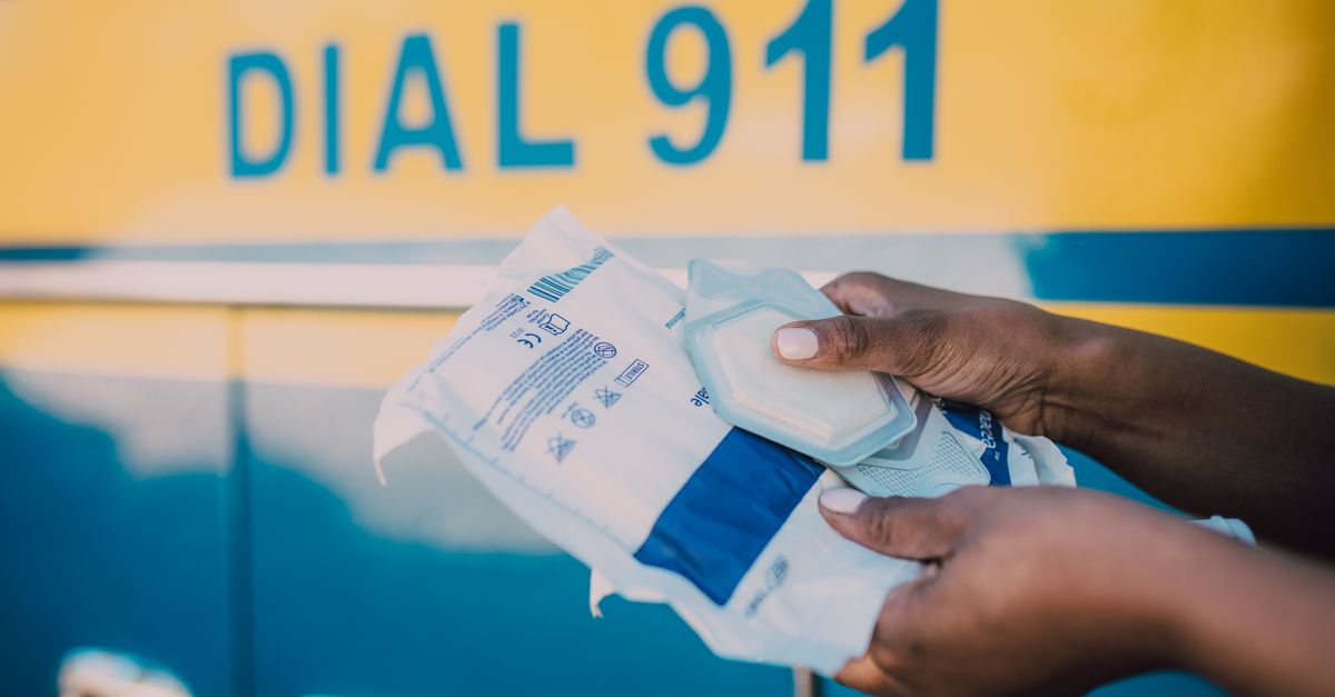 Hands holding medical supplies in front of an ambulance with 'Dial 911' sign.
