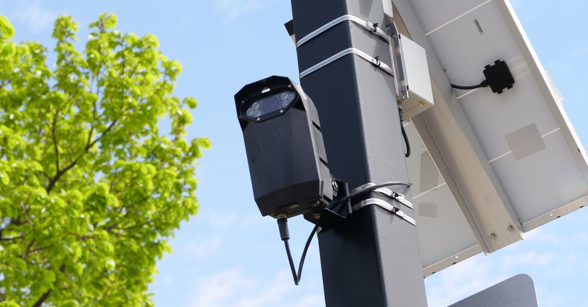 Urban surveillance camera mounted on pole with solar panel and green tree in view.