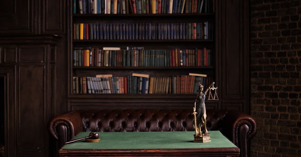 A classic study room with shelves of books, a gavel, and Lady Justice figurine on a green table.