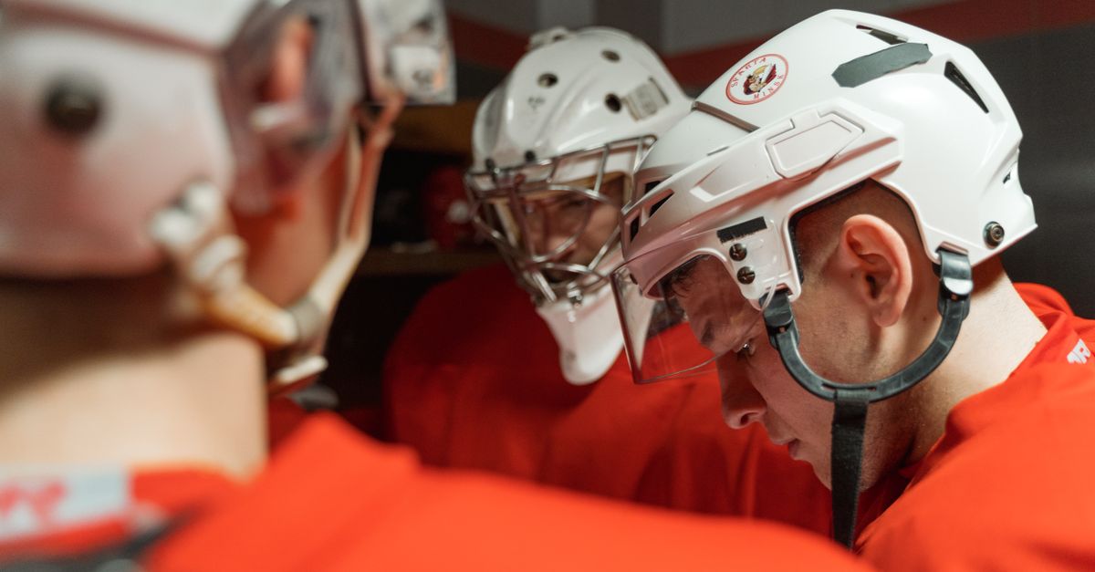 A group of male ice hockey players in helmets and uniforms huddled in a locker room preparing for a game.