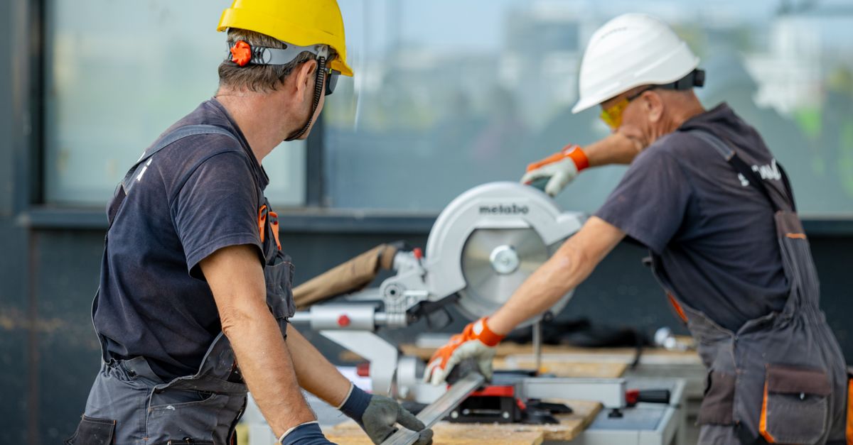Two construction workers cutting a metal beam with a circular saw, wearing safety gear on site.