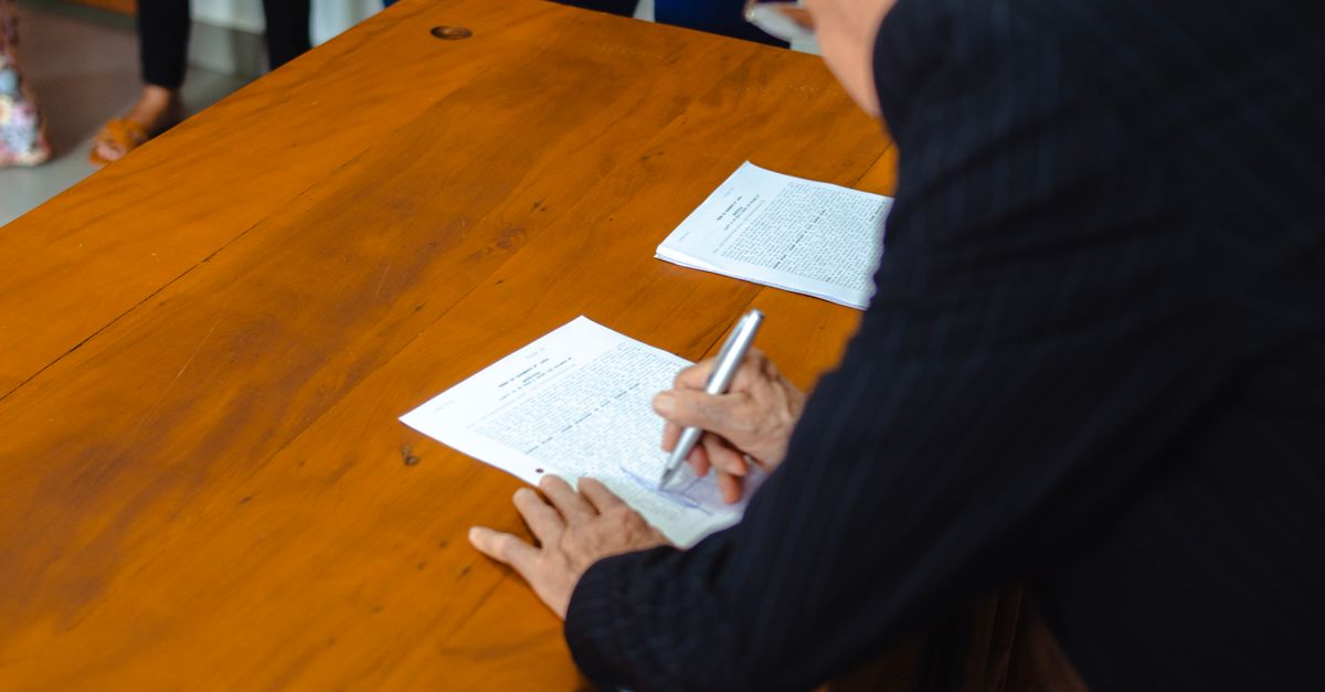 A close-up of a businessman signing official documents at a wooden desk.