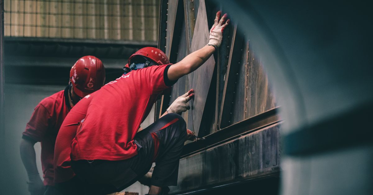 Two workers in red helmets assembling metal components inside a factory.