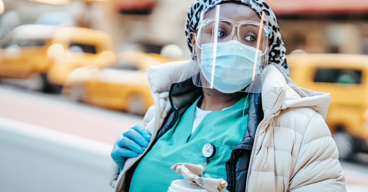 Thoughtful African American nurse in outerwear wearing medical mask face shield and gloves standing on busy street and looking away