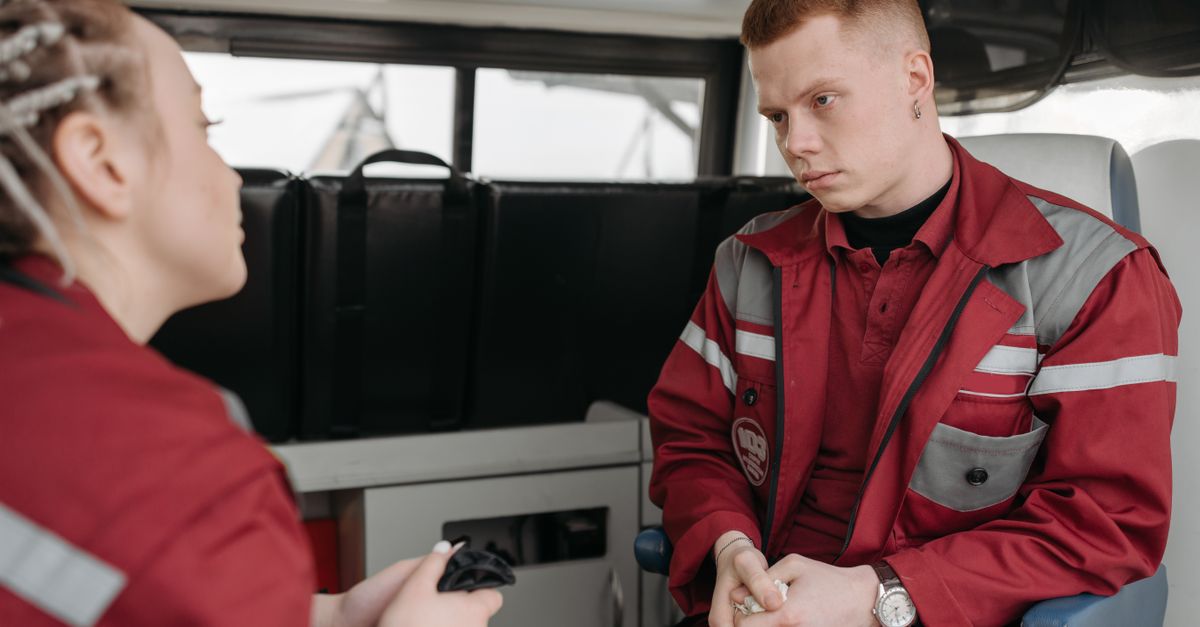 Two EMTs in burgundy uniforms engaged in serious conversation inside an ambulance.