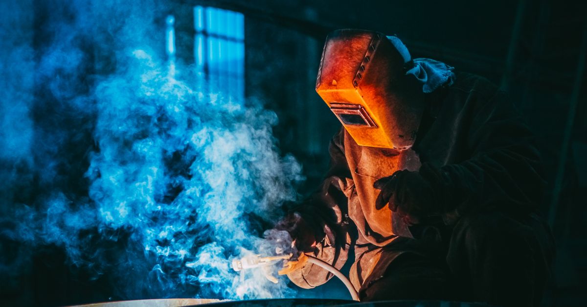 A welder in a workshop using safety gear to weld metal, surrounded by blue smoke and atmospheric lighting.